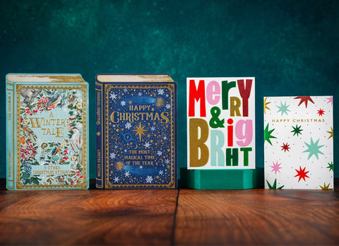 Four Christmas-themed greeting cards arranged on a wooden table against a dark teal textured background. The set includes a vintage-style illustrated book, a blue 'Happy Christmas' book, a colorful 'Merry & Bright' card, and a white 'Happy Christmas' card with star patterns.