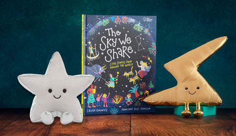 A children's book titled 'The Sky We Share' stands upright on a wooden surface between two plush star-shaped toys. The left toy is light gray and the right toy is metallic gold, both with embroidered smiling faces.