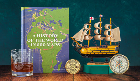 A book titled 'A History of the World in 500 Maps' stands on a wooden surface beside a model sailing ship, a compass, a brass calendar paperweight, and a glass with amber liquid, all set against a dark teal background.