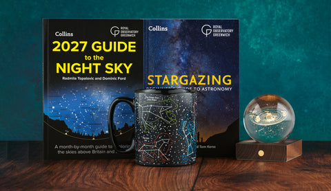 A set of astronomy-themed products displayed on a wooden surface against a teal textured background: a hardcover book titled '2027 Guide to the Night Sky,' a hardcover book titled 'Stargazing: A Guide to Astronomy,' a matching black ceramic mug with a starry night sky print, and a glass planetarium dome on a wooden base.