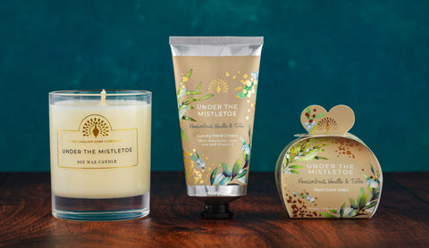 Three scented products from Under the Mistletoe by The Scented Candle Co. displayed together on a dark wooden surface against a deep teal background: a glass jar candle, a squeeze tube, and a heart-shaped box.