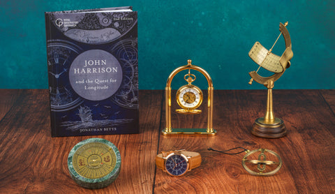 A styled tabletop scene featuring a book titled "John Harrison and the Quest for Longitude" by Jonathan Betts, alongside several clocks and navigational instruments on a wooden surface against a teal background.