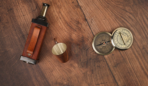 A vintage-style brass telescope with a brown leather strap, a small copper tumbler, and two brass compasses arranged on a dark wood tabletop.