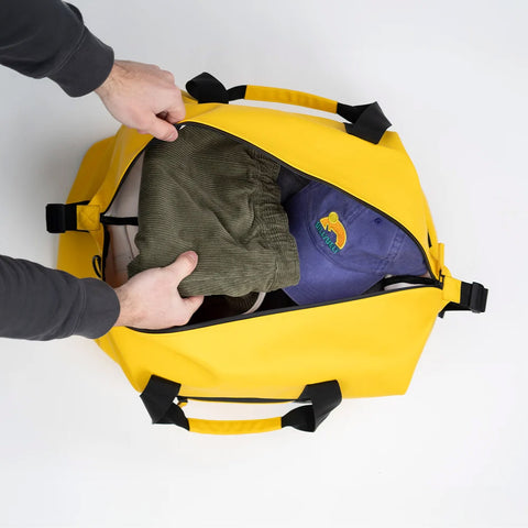 A bright yellow duffel bag with black handles and zippers, shown from above as a person's hands open the top zipper 