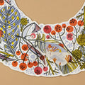 A close-up of a decorative illustrated advent wreath laid flat on a tan background. It features a whimsical winter botanical print with red berries, yellow flowers, blue leaves, and a small snowman motif.