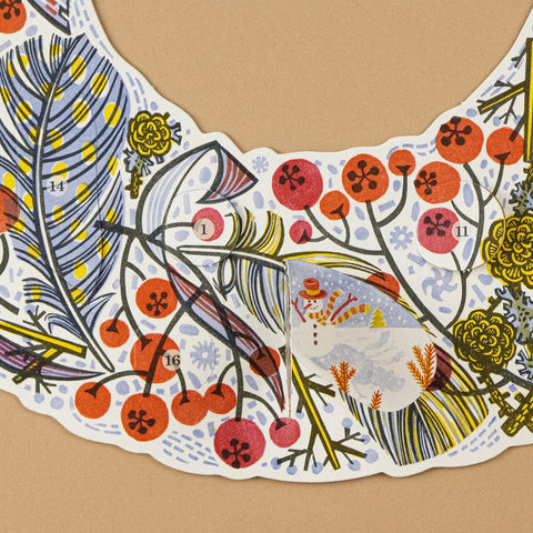 A close-up of a decorative illustrated advent wreath laid flat on a tan background. It features a whimsical winter botanical print with red berries, yellow flowers, blue leaves, and a small snowman motif.
