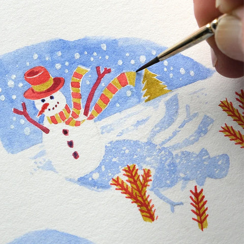 A close-up of a hand painting a whimsical snowman scene on textured white paper, using a fine paintbrush to add details to a watercolor illustration with blue, red, yellow, and white tones.