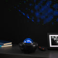 A black tabletop night light projector with a glowing blue globe design sits on a dark surface, casting blue and green starry light patterns onto a black wall. A remote control, stacked books, and a framed picture are visible nearby in a dim room setting.