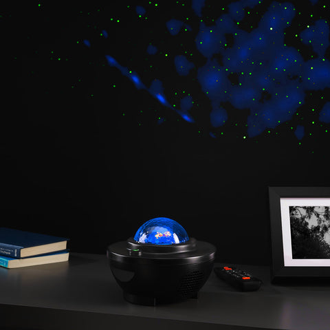 A black tabletop night light projector with a glowing blue globe design sits on a dark surface, casting blue and green starry light patterns onto a black wall. A remote control, stacked books, and a framed picture are visible nearby in a dim room setting.