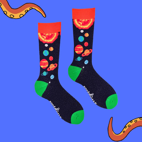 A pair of crew socks with a black space-themed print featuring colorful planets, stars, and a red-orange sun motif on the cuff, shown against a bright blue background with decorative orange tentacle graphics in the corners.
