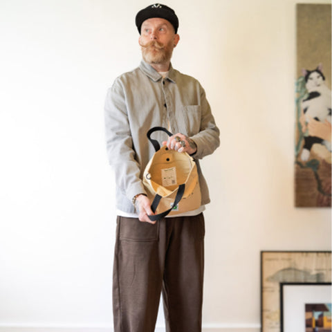 Man holding open a beige tote bag indoors with a light-coloured wall and framed picture in the background