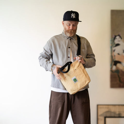 Man holding a beige bag with a Cora + Spink logo in a room with a painting on the wall.