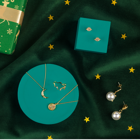 Gold crescent moon necklace and gold jewelled pendant necklace on green background, surrounded by constellation gold ring, gold Saturn earrings and gold pearl drop earrings