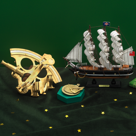 Gold sextant replica, golden replica sundial, Cutty Sark model ship and The Camden Watch Company Cutty Sark Royal Museums Greenwich watch arranged on a green starry setting with wrapped Christmas gifts