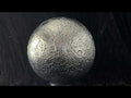 Miniature stainless steel replica of the Moon rotating to show all areas of the surface. Craters and topographical details are accurate according to NASA imagery. Light reflects off the silver surface, illuminating the moon sculpture against a black curtain backdrop. 