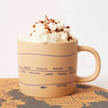 Beige mug with blue text and design, filled with hot chocolate and whipped cream, on a map.