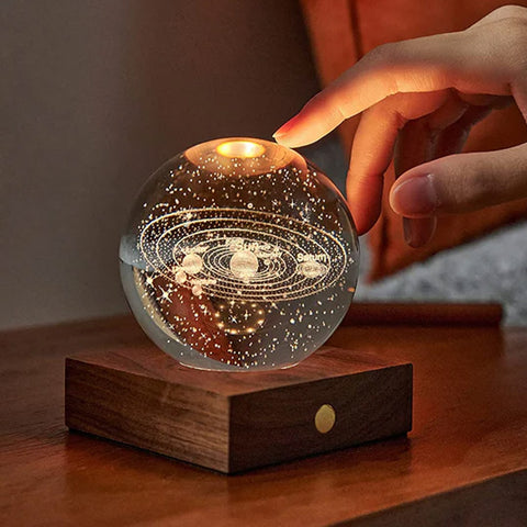 A hand touching a 3D laser-engraved design of the solar system in a crystal ball on a wooden base.