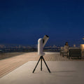 Telescope on a tripod with a person sitting on a rooftop at night, looking at the stars.