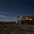 Telescope on a tripod in the foreground with a camper van and two people under a starry night sky