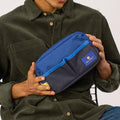 A person in an olive green corduroy shirt holds a blue and navy zippered pouch with a visible logo patch.