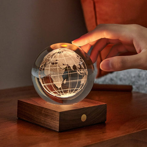 Hand touching lit 3D engraved globe design in a crystal ball on a wooden stand.