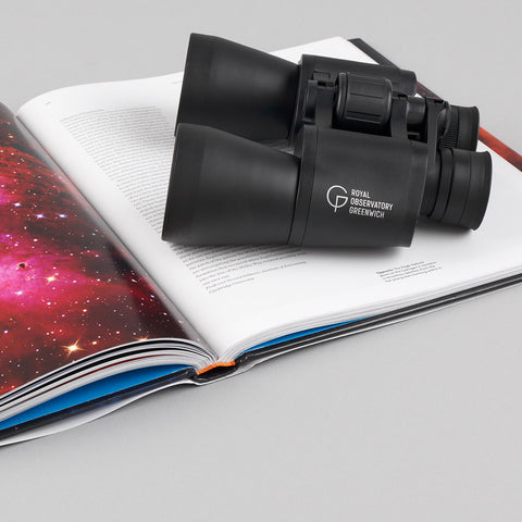 Black binoculars with a white Royal Observatory Greenwich logo, placed on an open astronomy book.