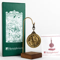 Small brass Astrolabe Astrolabio hanging from stand next to green box with renaissance style illustration.